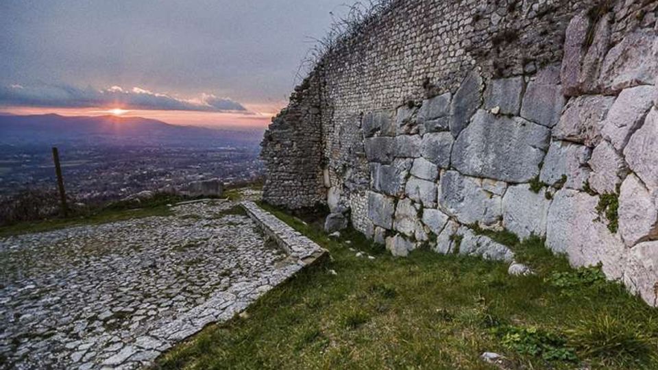 Spiazzo in pietra acciottolata accanto a un alto muro di fortificazione in pietra, con tratti di muratura antica e blocchi irregolari. Sullo sfondo si vede una vasta valle urbana al tramonto, con cielo nuvoloso e orizzonte montuoso.