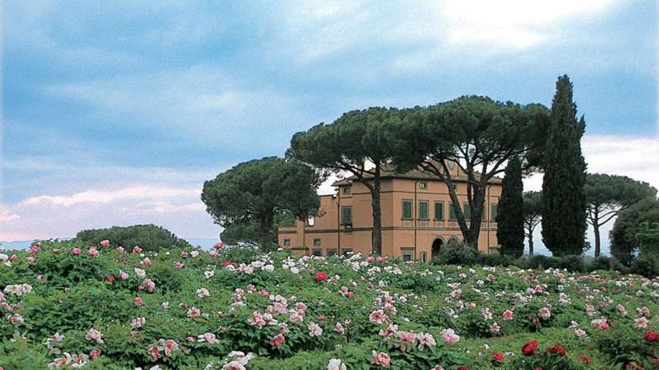 Villa di campagna color ocra con finestre e un portico ad archi, circondata da alti pini e cipressi. In primo piano si estende un ampio campo di fiori rosa, bianchi e rossi sotto un cielo nuvoloso.