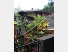 Casa con veranda in legno e tetto spiovente, costruita su un terrazzamento con muri in pietra. In primo piano una scalinata con ringhiera in legno e vasi di piante, circondata da vegetazione e alberi.