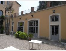 Cortile con ghiaia davanti a un edificio basso color ocra, con grandi porte e finestre ad arco incorniciate di bianco e infissi grigi. Sulla facciata ci sono lampade a parete, una ringhiera sul tetto e alcune piante e arbusti in vaso vicino all’ingresso.