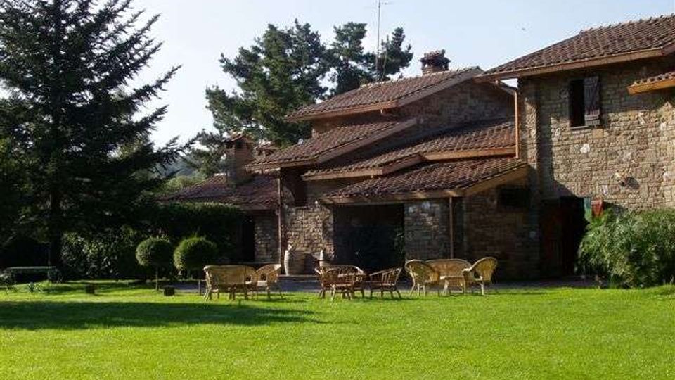 Edificio rurale in pietra con tetti in tegole rosse e portico, affacciato su un ampio prato verde con sedie e tavolini da giardino. Alberi alti e cespugli delimitano l’area esterna sullo sfondo.