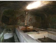 Interno di una grotta o struttura in pietra con vasche rettangolari in muratura piene d’acqua e una piccola fonte centrale che sgorga dal muro. Una luce dall’alto illumina parzialmente le pareti scure e umide.