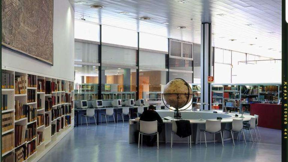 Interno di una biblioteca moderna con scaffali di libri lungo le pareti, postazioni con computer e tavoli di studio. Ampie vetrate e soffitto a pannelli illuminano lo spazio, con un grande globo su un tavolo centrale.