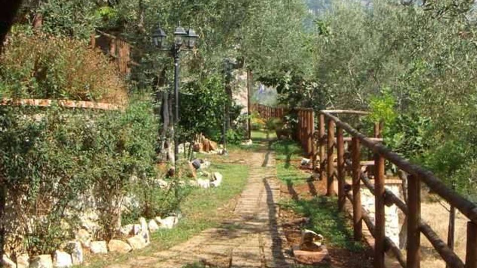 Sentiero in pietra e terra che attraversa un giardino rurale con ulivi e vegetazione bassa, fiancheggiato da una staccionata in legno a destra e muretti con rocce e lampioni a sinistra. Sullo sfondo si intravedono colline sotto un cielo sereno.