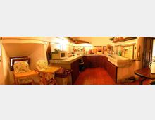 Cucina rustica in mansarda con soffitto spiovente, pavimento in cotto e mobili in legno scuro disposti a L. Piano di lavoro in pietra con lavello e mensole illuminate, piccoli elettrodomestici e due seggioloni con vassoi vicino a una finestra bassa.