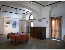 Interno di una piccola sala espositiva con soffitto a volte e archi bianchi, illuminazione a binari e pavimento in pietra. Al centro c’è un tavolo con tovaglia rossa, mentre lungo le pareti si vedono pannelli informativi, una grande cassettiera in legno e una finestra ad arco con tenda a righe.
