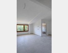Stanza interna non arredata con pareti bianche e pavimento in piastrelle chiare, illuminata da una grande finestra con infisso in legno e vista sul verde esterno. A destra si vede un’apertura verso un bagno piastrellato, con impianti elettrici a vista e soffitto inclinato.