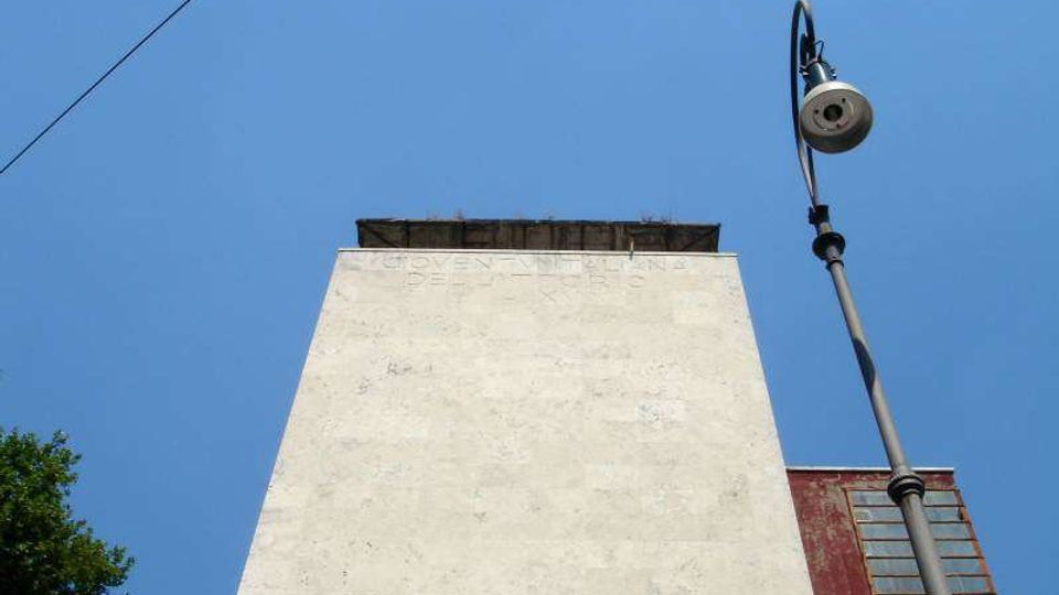 Vista dal basso di una torre rettangolare in muratura chiara con sommità sporgente, contro un cielo azzurro. A destra si vede un lampione alto e, sullo sfondo, una facciata rossa con finestre verticali.