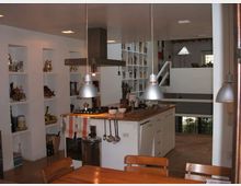 Interno di una cucina moderna a pianta aperta con isola centrale in legno, cappa a soffitto e lampade a sospensione in metallo. Pareti con nicchie e scaffalature piene di oggetti, tavolo in legno in primo piano e soppalco con ringhiera che affaccia sull’ambiente.