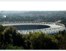 Vista panoramica di un grande stadio ovale con struttura in travi bianche e copertura a membrane, circondato da alberi e aree verdi. Sullo sfondo si vede la città con edifici bassi e strade in lontananza sotto un cielo chiaro.