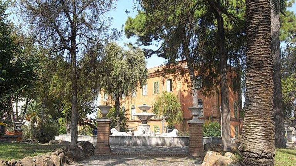 Vialetto in pietra che attraversa un parco alberato con palme e pini, delimitato da muretti di roccia. Sullo sfondo si vede un ingresso con pilastri e una villa storica color ocra tra la vegetazione.