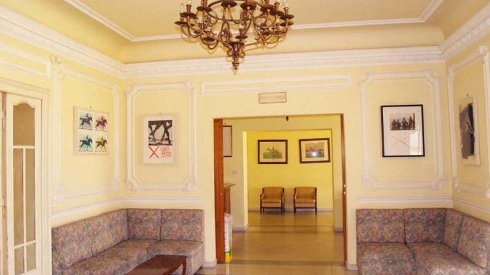 Salotto o area d’attesa in stile classico con pareti gialle decorate da cornici in gesso, lampadario centrale e divani angolari con tavolino. Un’ampia apertura conduce a una stanza adiacente con sedie e quadri, su pavimento a scacchi.
