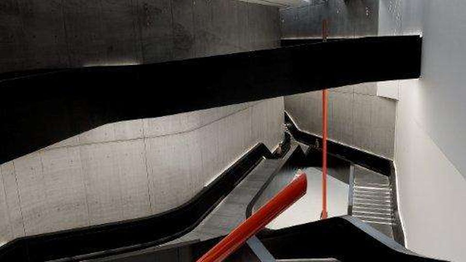 Interno di una scala moderna in cemento a più rampe con parapetti neri curvi, corrimano rosso e gradini in griglia metallica. In alto un lucernario longitudinale illumina l’ampio vano verticale.