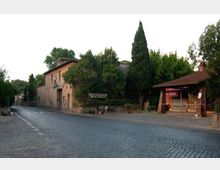 Strada lastricata in pietra in un’area rurale, con un edificio in muratura stile cascina e vegetazione alta sullo sfondo. A destra si vede una piccola pensilina in legno e mattoni con cartelli, affacciata sulla strada.