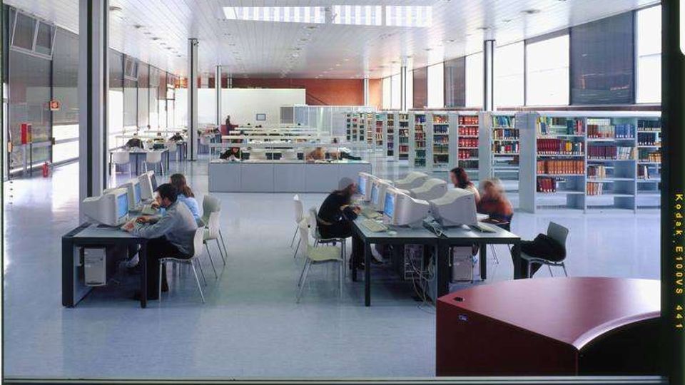 Ampia sala di biblioteca moderna con soffitto alto a pannelli e grandi finestre laterali che illuminano lo spazio. Sono presenti tavoli con postazioni computer, sedie e lunghe file di scaffali con libri disposti in modo ordinato.