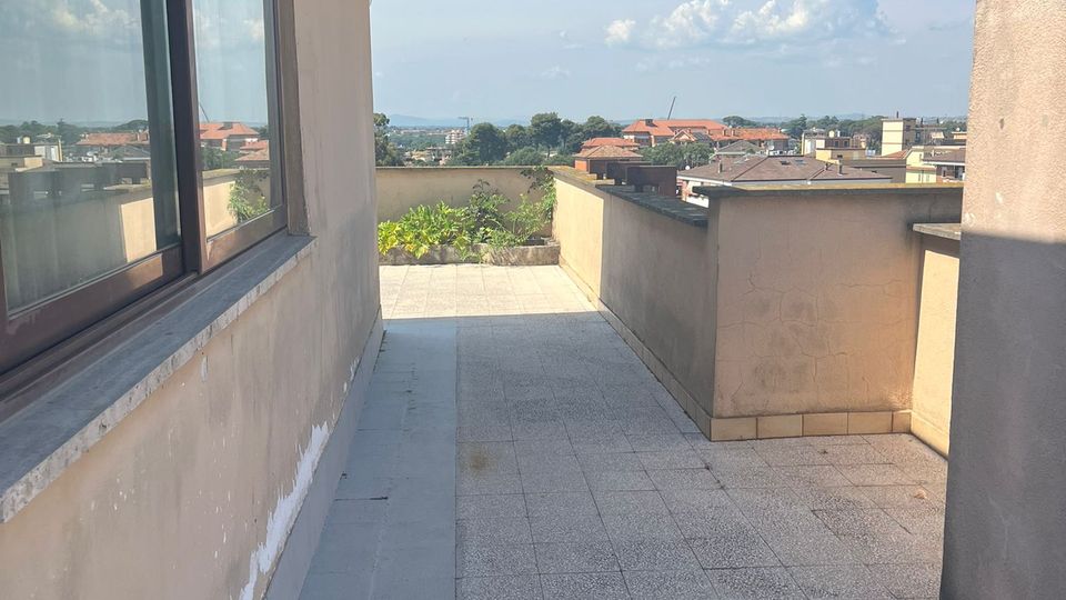 Terrazza sul tetto con pavimento piastrellato e parapetti in muratura, affiancata da una parete con grandi finestre e sporgenza del tetto. In fondo si vede una fioriera con vegetazione e un panorama urbano con tetti e cielo azzurro.