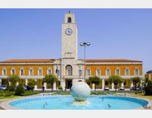 Edificio pubblico simmetrico in stile razionalista con facciata giallo ocra e una torre centrale in pietra con orologio. In primo piano si vede una grande fontana circolare con una sfera al centro, circondata da aiuole e alberi in una piazza.