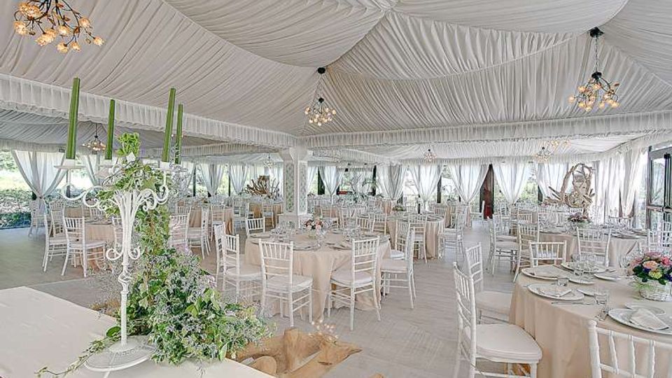 Ampia sala ricevimenti allestita con tavoli rotondi e sedie bianche, illuminata da lampadari e con soffitto completamente drappeggiato in tessuto bianco. Grandi tende bianche lungo le pareti e composizioni floreali sui tavoli definiscono l’ambiente.