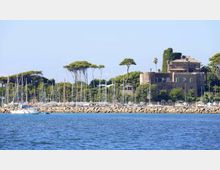 Veduta di un porto turistico con numerose barche a vela ormeggiate dietro una diga frangiflutti in pietra, con il mare in primo piano. Sullo sfondo si vede una grande villa in muratura tra pini marittimi e vegetazione costiera.