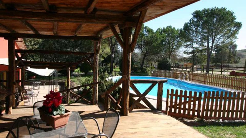 Veranda in legno coperta con tavoli e sedie da esterno, affacciata su una piscina rettangolare recintata con staccionata in legno. Sullo sfondo si vedono prato e alberi alti in un’area verde.
