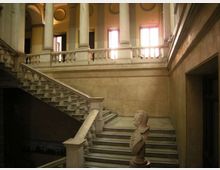 Interno di un edificio storico con ampia scalinata in pietra e balaustra, che conduce a un loggiato con alte colonne e arcate. Grandi finestre sul piano superiore illuminano pareti intonacate e un busto su piedistallo alla base della scala.