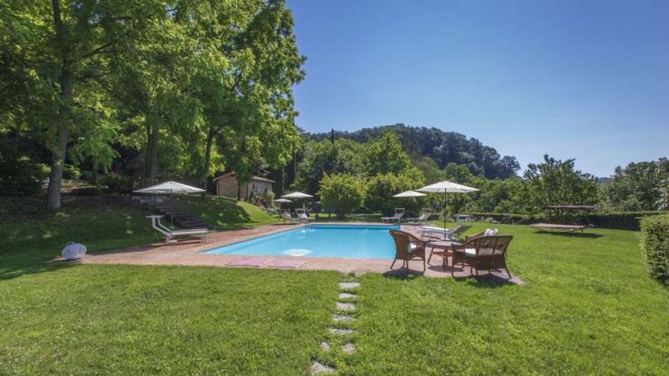 Piscina all’aperto rettangolare circondata da un ampio prato verde con sedie a sdraio, tavoli e ombrelloni. Sullo sfondo si vedono alberi alti, una collina boscosa e alcune piccole strutture in muratura ai margini del giardino.