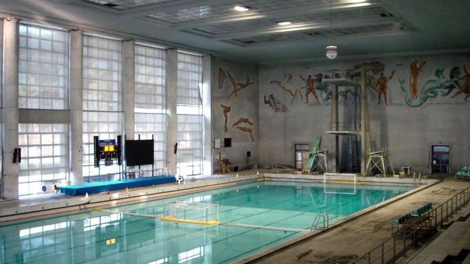 Piscina coperta con grande vasca rettangolare e acqua turchese, illuminata da ampie finestre a tutta altezza su un lato. Pareti decorate con murales di figure atletiche e trampolino alto con piattaforme, con tribune e corrimano lungo il bordo vasca.