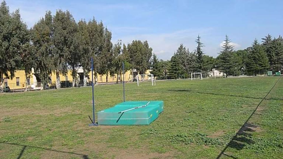 Campo sportivo all’aperto con area verde e una pedana per salto in alto con materasso verde e due aste verticali. Sullo sfondo si vedono alberi, alcune porte da calcio e edifici bassi lungo il perimetro.