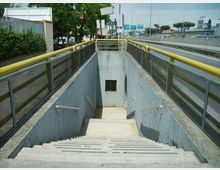 Scala in cemento che scende a un sottopassaggio pedonale, con corrimano laterali e parapetti in rete metallica con fascia gialla. Ai lati si vede una strada a più corsie con guardrail e segnaletica, con alberi e edifici sullo sfondo.