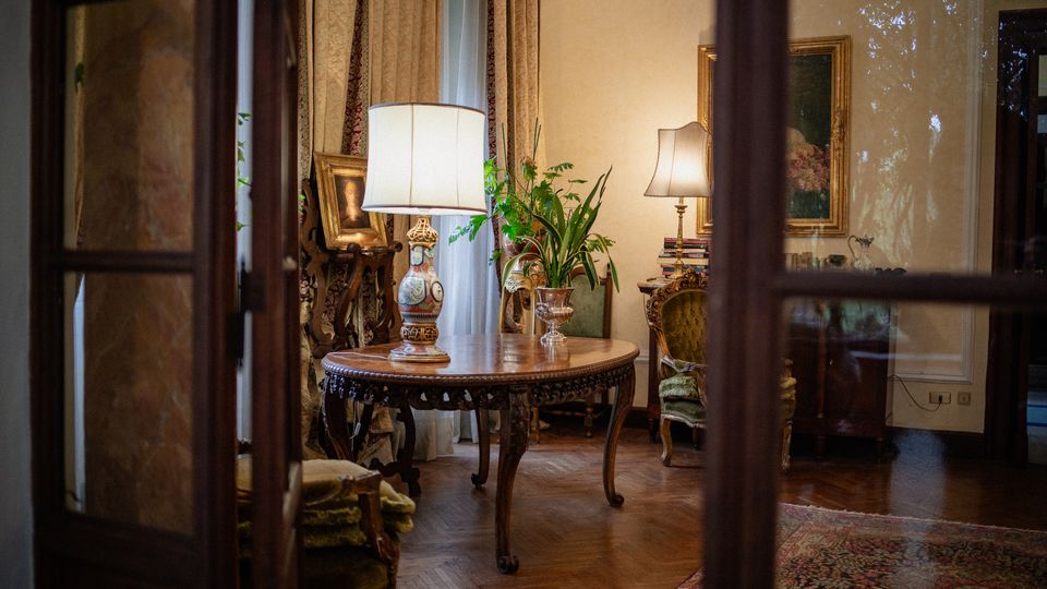Interno di un salotto classico visto attraverso una porta a vetri, con pavimento in parquet e tappeto. Al centro c’è un tavolo rotondo intagliato con lampada, circondato da poltrone imbottite, quadri incorniciati, tende pesanti e piante vicino alla finestra.