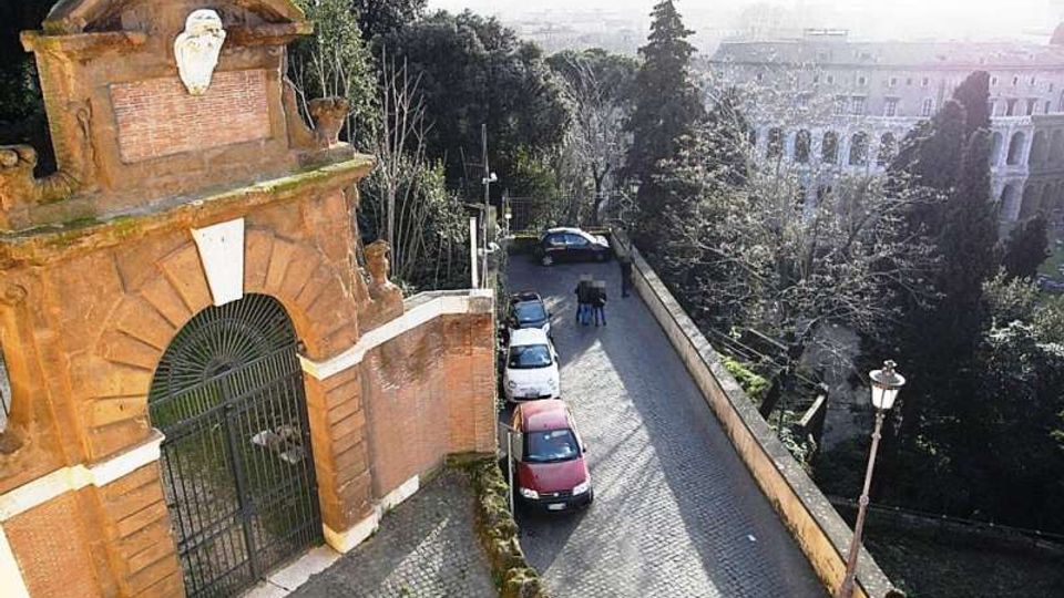 Strada in sampietrini in discesa accanto a un alto muro, con auto parcheggiate lungo il lato e lampione in primo piano. A sinistra si vede un grande portale in mattoni con arco e cancello in ferro, circondato da alberi e con vista su edifici sullo sfondo.