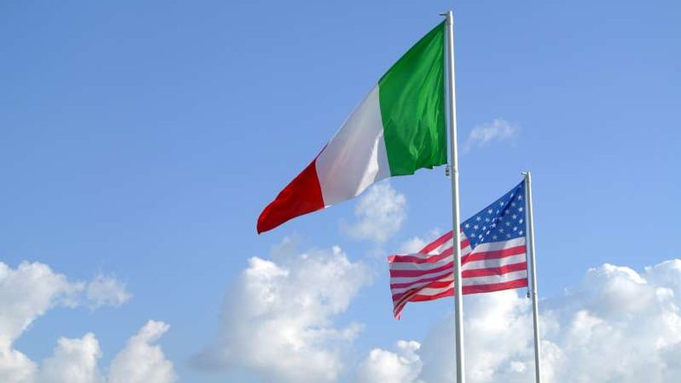 Due pennoni con la bandiera italiana e la bandiera degli Stati Uniti che sventolano all’aperto contro un cielo azzurro con nuvole bianche.