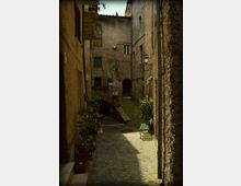 Vicolo stretto in un borgo storico con pavimentazione in ciottoli, fiancheggiato da edifici in pietra con persiane e lampioni a muro. In fondo si vede un piccolo ponte ad arco e una rampa con vasi di piante; sul lato destro c’è una panchina verde vicino alla parete.