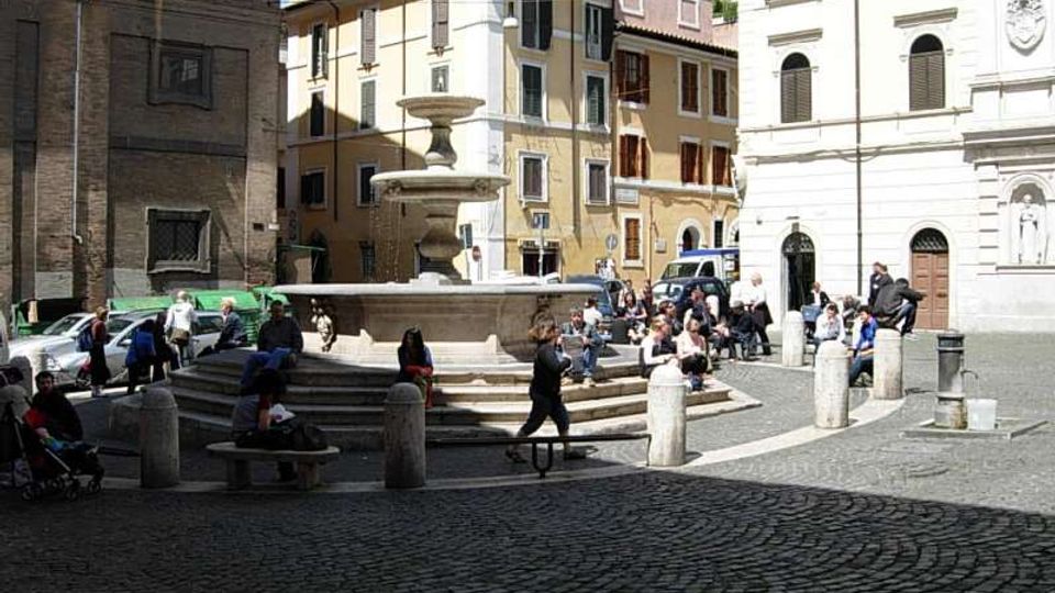 Piazza lastricata in sampietrini con una grande fontana in pietra al centro e gradini tutt’intorno. Sullo sfondo si vedono edifici storici a più piani con persiane e facciate chiare, oltre a una facciata di chiesa e alcuni dissuasori in pietra lungo il bordo della piazza.
