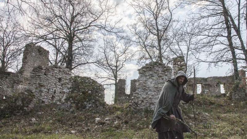 Rovine di un edificio in pietra con muri parzialmente crollati su un pendio erboso, circondato da alberi spogli. Sullo sfondo si vede un cielo nuvoloso sopra i resti delle strutture.