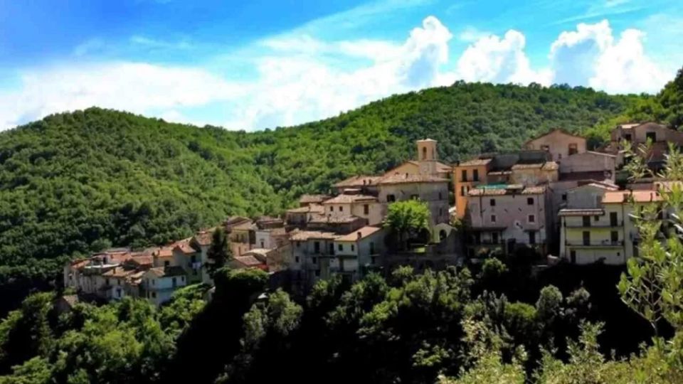 Veduta panoramica di un borgo collinare con case in pietra dai tetti in coppi, raggruppate su un pendio e circondate da fitti boschi. Sullo sfondo si vedono colline verdi e un cielo azzurro con nuvole.
