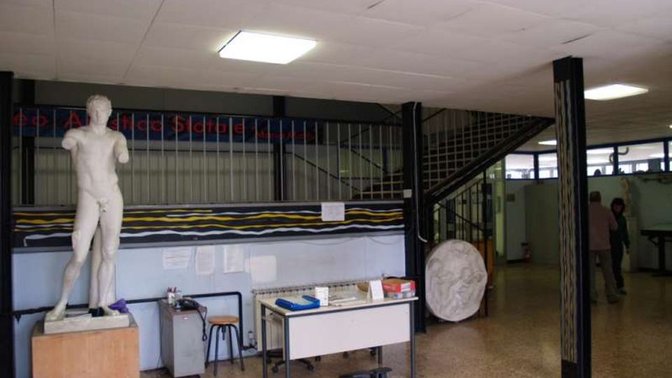 Interno di un laboratorio scolastico o artistico con pavimento in graniglia, soffitto a pannelli e luci al neon, con scrivanie e attrezzature. A sinistra c’è una statua classica in gesso su basamento, mentre sullo sfondo si vedono una scala metallica e una ringhiera che affacciano su un soppalco.