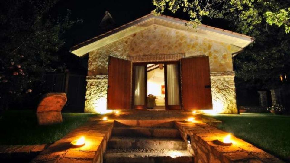 Piccolo edificio in pietra illuminato di notte, con tetto a due falde e grandi porte in legno aperte su un ingresso. Un breve vialetto con gradini in pietra e luci a terra conduce dalla zona erbosa fino alla soglia, circondato da alberi.