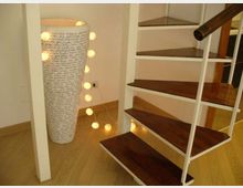 Interno domestico con scala a chiocciola in metallo bianco e gradini in legno scuro su pavimento in parquet. Accanto alla scala c’è una lampada cilindrica alta con scritte e una catena di luci a sfera appoggiata a terra.
