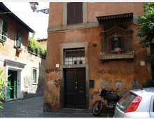 Facciata di un edificio color ocra su una stretta strada acciottolata, con portone nero, finestra con inferriata e graffiti sui muri. A destra sporge un piccolo balconcino decorato con edicola e lampada, con una moto parcheggiata e parte di un’auto in primo piano.