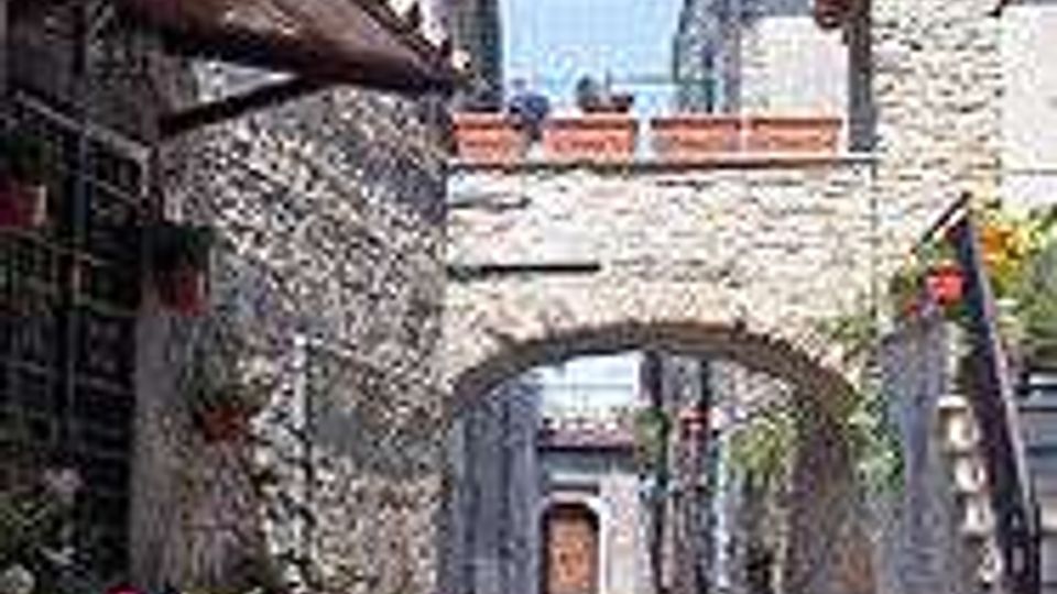 Vicolo lastricato in pietra tra edifici in muratura, con arco in pietra che collega le case e una scalinata laterale con ringhiera. Ai lati si vedono finestre, persiane e vasi di fiori, in un contesto di borgo storico all’aperto.