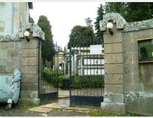 Ingresso di un cimitero con cancello in ferro battuto tra due pilastri in pietra con lampade sferiche. Sullo sfondo si vedono cappelle e tombe tra alberi e siepi, con un percorso interno in ghiaia.
