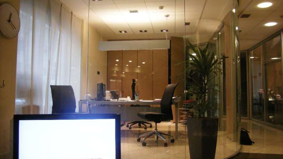 Ufficio moderno con pareti vetrate curve e pavimento in piastrelle chiare, arredato con scrivania centrale, sedie su ruote e un grande vaso con pianta. Sullo sfondo si vedono pannelli in legno, illuminazione a soffitto e, in primo piano, uno schermo acceso vicino a una finestra con tende bianche.