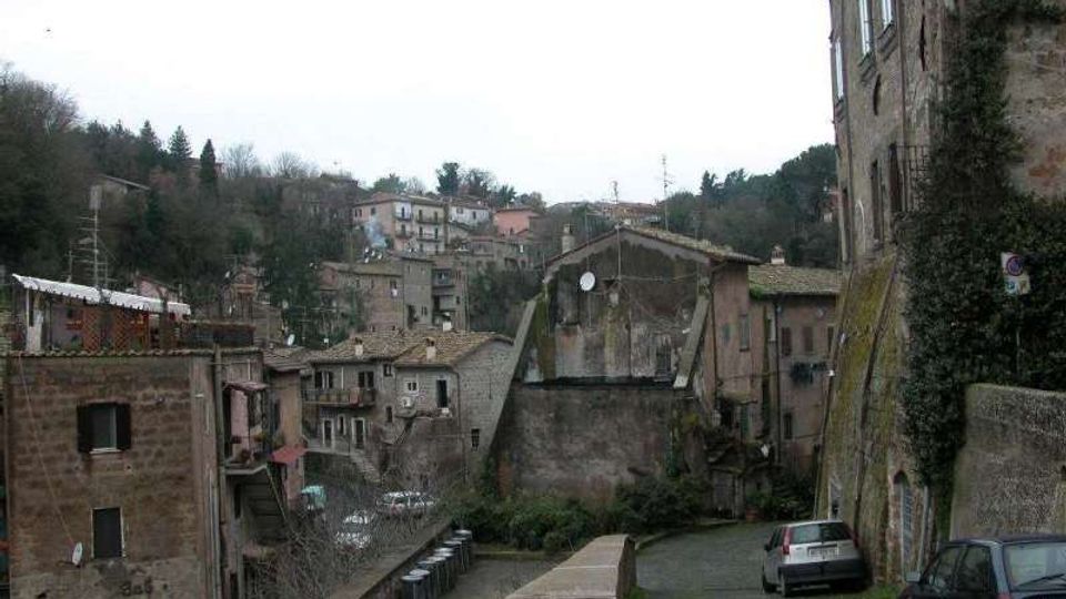Vista di un borgo collinare con case in pietra e tetti in coppi, disposto su più livelli lungo il pendio. In primo piano si vede una strada stretta con un muretto in pietra e alcune auto parcheggiate, mentre sullo sfondo ci sono alberi e abitazioni sparse sulla collina.