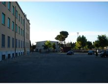 Ampio piazzale asfaltato con alcuni parcheggi e pochi veicoli, affiancato a sinistra da un edificio a più piani con file di finestre. Sullo sfondo si vedono alberi, una struttura bassa tipo capannone e un’area recintata sotto un cielo sereno.