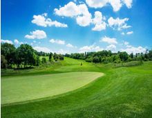 Campo da golf con ampio green in primo piano e bandierina centrale, circondato da colline erbose e alberi. Sullo sfondo cielo azzurro con nuvole sparse.