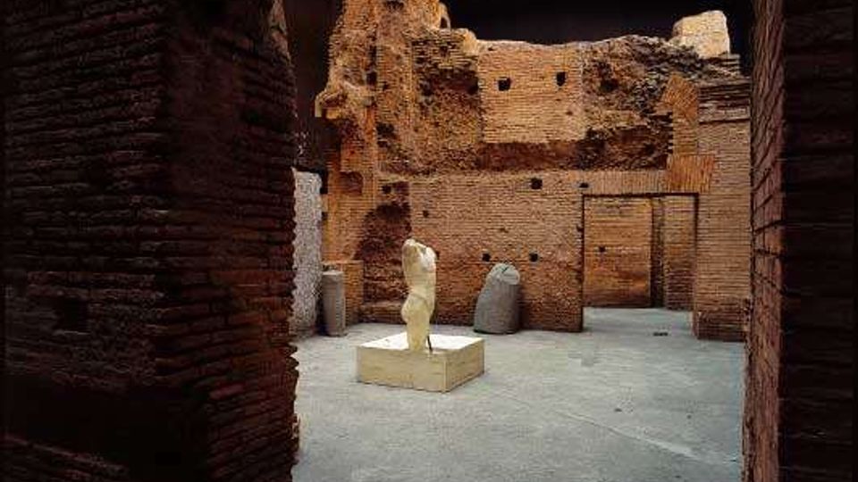 Interno di un’area archeologica coperta, con pavimento in cemento e pareti in mattoni antichi parzialmente crollate. Al centro si trova una piccola statua su basamento, sotto una grande copertura con travi metalliche e pannelli traslucidi.