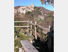 Vista panoramica di un borgo medievale arroccato su una collina rocciosa, con case in pietra addossate tra loro e una torre in cima. In primo piano si vede un sentiero con staccionata in legno e vegetazione, con colline boscose sullo sfondo sotto un cielo sereno.
