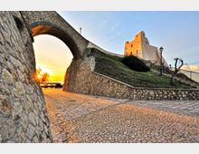 Veduta di una fortificazione su una collina, con mura e arco in pietra sopra una strada acciottolata che conduce a un edificio storico. Sullo sfondo si vedono scale con ringhiera, lampioni e il cielo al tramonto.