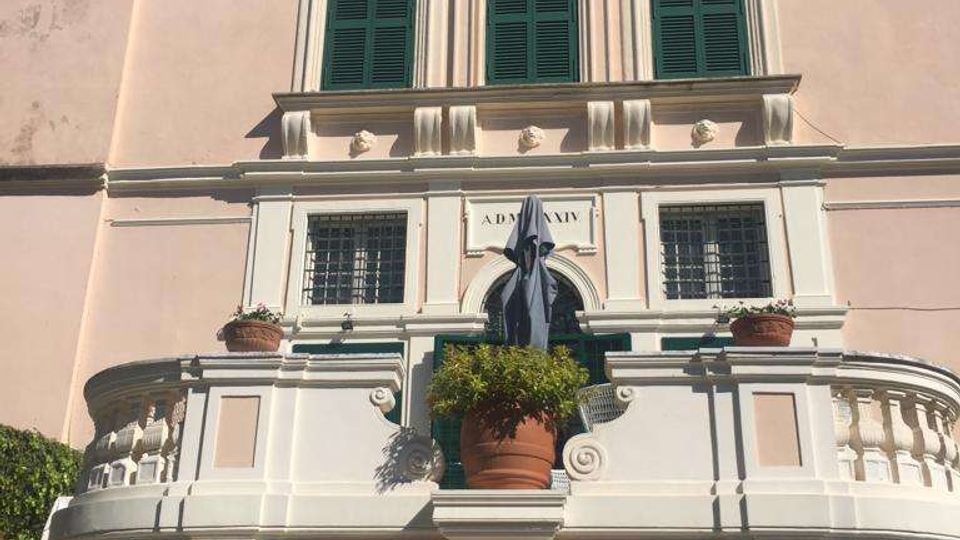 Facciata di un edificio storico a più piani con intonaco chiaro, finestre con persiane verdi e cornici decorative. Al centro si vede un balcone semicircolare con balaustra, vaso e ombrellone chiuso sopra un grande portone con inferriata in ferro battuto.